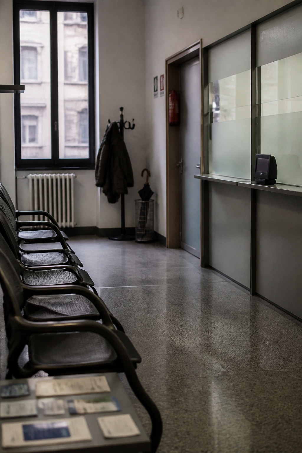 Sala d'attesa di un patronato italiano, banco in vetro acidato, distributore di numeri e scaffale di moduli a faccia in giù
